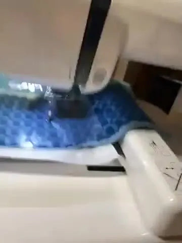 Close-up of the Janome Memory Craft 230E stitching a green design onto a blue checkered towel under clear water-soluble stabilizer.