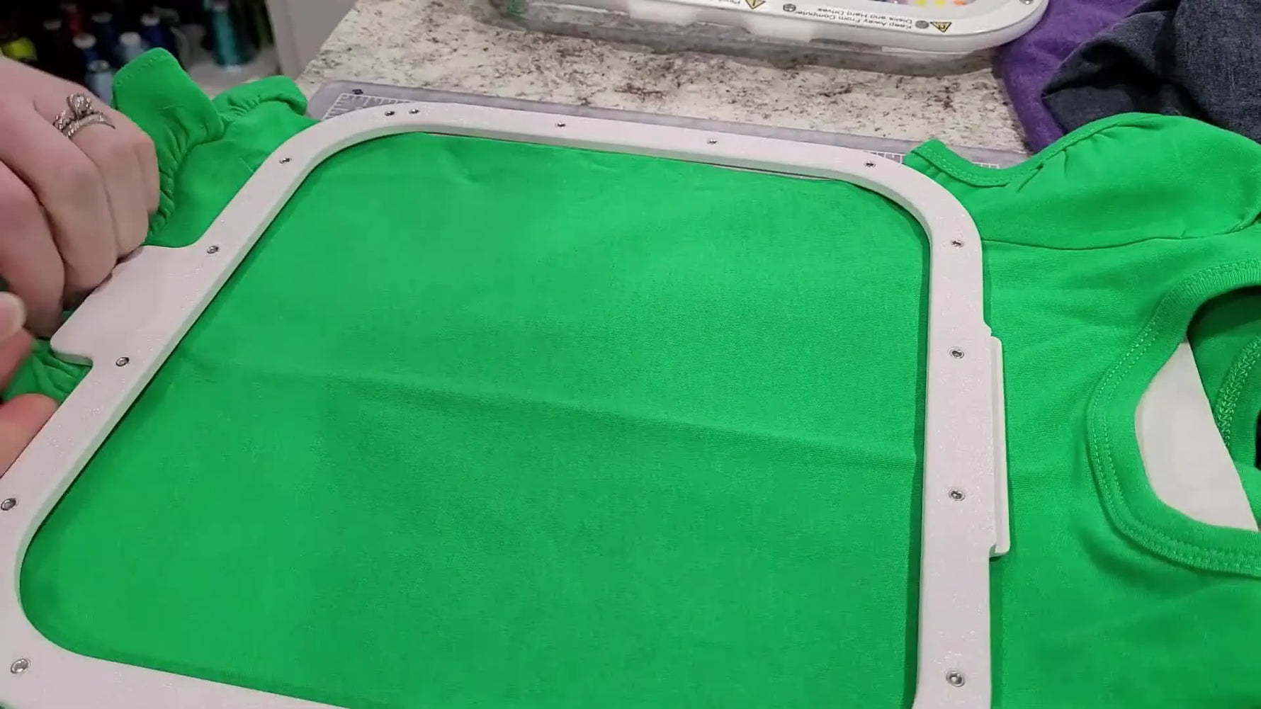 Hands snap a magnetic hoop onto a green children’s T‑shirt while checking center alignment marks before stitching on a Brother PR1000e.