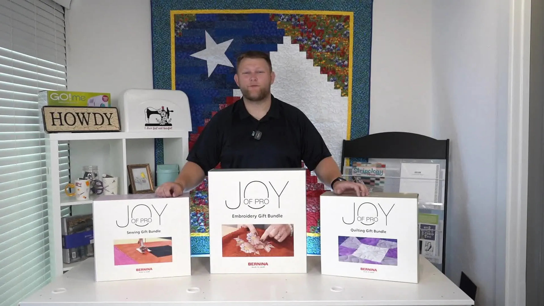 A presenter stands behind three large “Joy of Pro” gift boxes laid out on a table, previewing the embroidery, quilting, and sewing bundles.