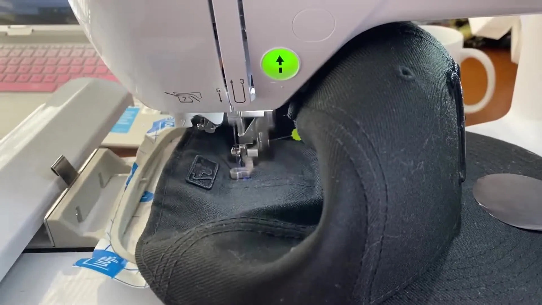 Close-up of a single-needle embroidery machine stitching blue name text onto the back arch of a fitted hat.