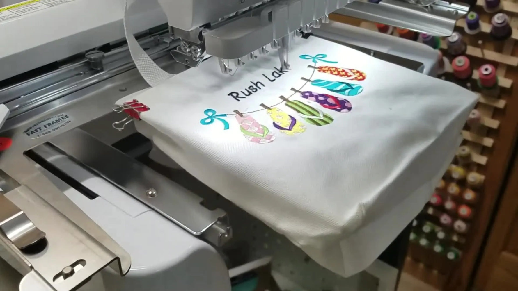 A multi-needle embroidery machine stitches a colorful design onto a tote bag mounted on a Fast Frames setup.