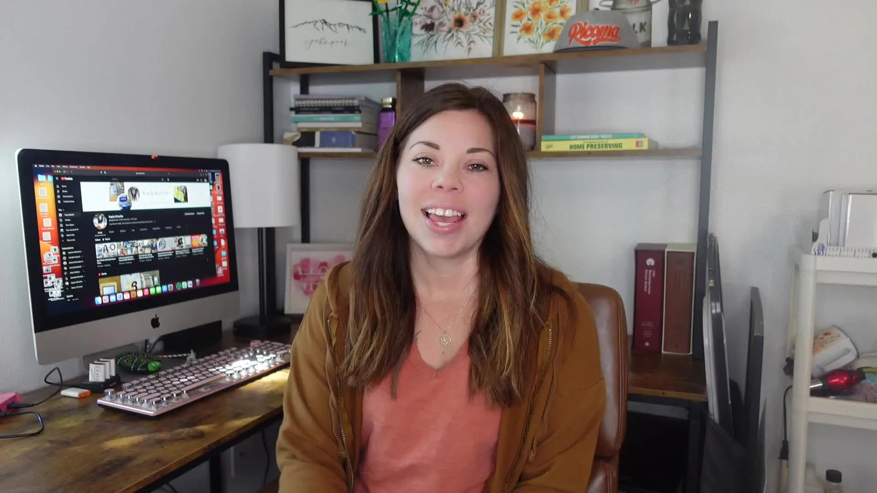 Kayla greeting viewers from her home office, with a computer showing a video editor and decor shelves behind her.