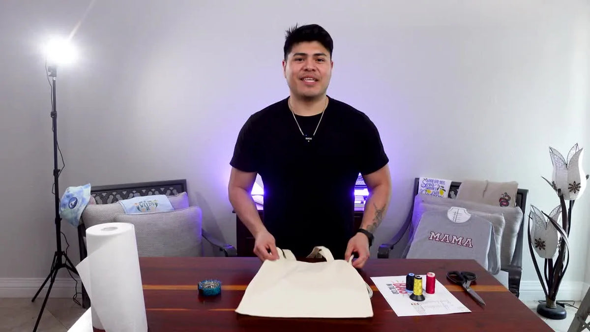 Man holding up a blank tote bag, smiling, with embroidery machine and supplies in the background.