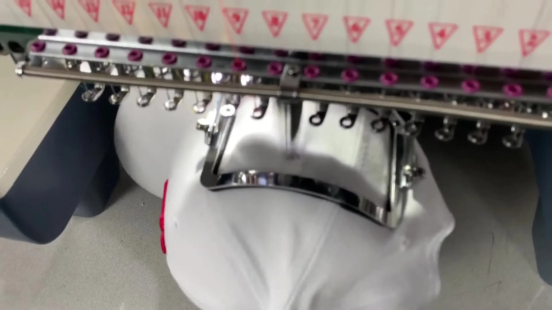 Close-up of the YunFu machine stitching a small design onto the side of a cap held firmly in the stainless Pocket/Cuff Device clamp.
