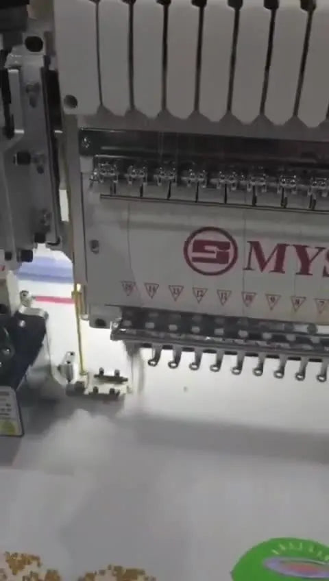 Close-up of the bead device actively stitching a curved line of gold beads onto fabric on a MYSEW 15-needle machine.