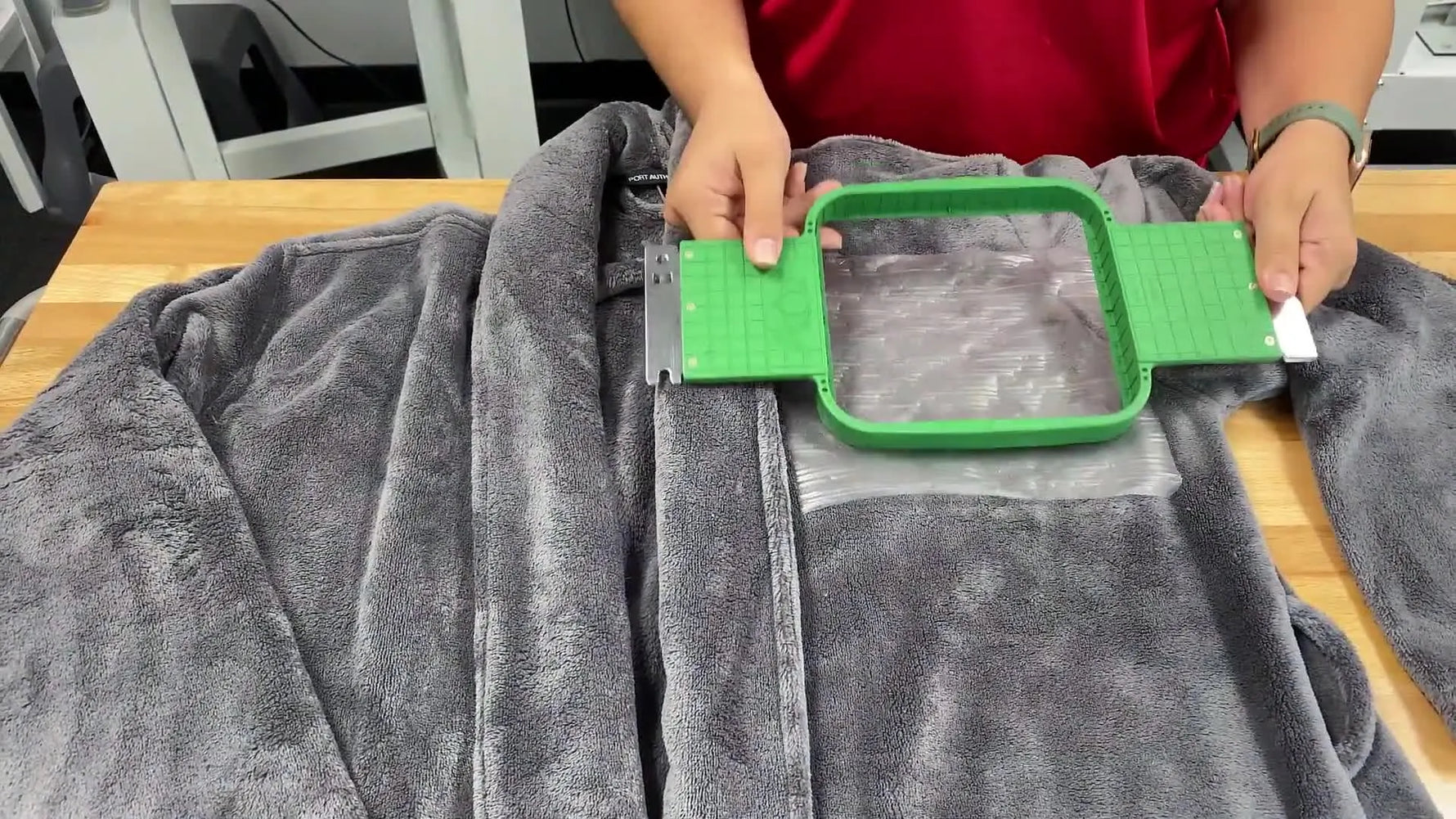 Hands press the green-handled Allied GridLock hoop onto a plush microfleece robe with topping in place, ready for a clean monogram sew-out.
