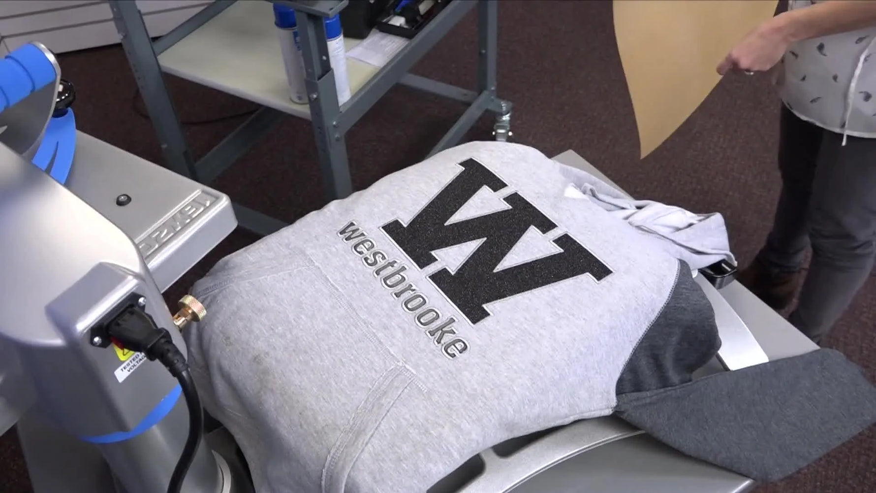 A presenter holds up a finished grey hoodie featuring a black mixed-media appliqué design with crisp white embroidery outlines.