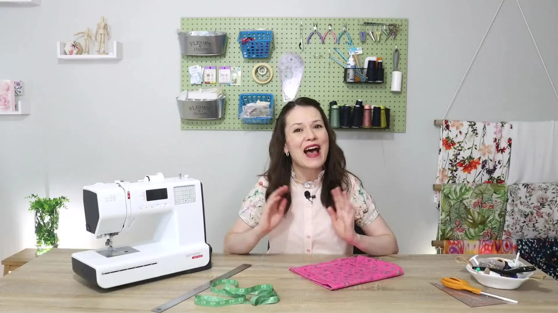 A woman in a floral blouse smiles at the camera, with a sewing machine and sewing supplies on a wooden desk in front of her. A pegboard with sewing tools is visible behind her.
