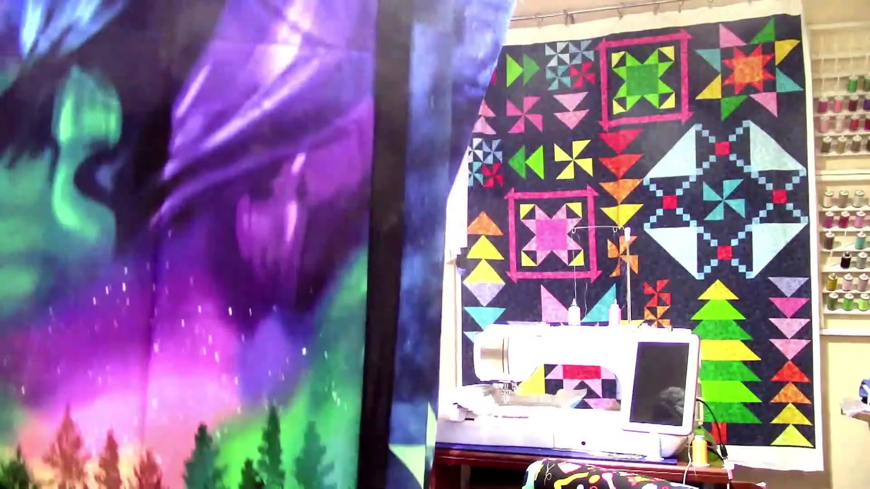 The host holds up a partially finished quilt top with star blocks and a northern-lights panel, showing real project progress in a busy sewing studio.