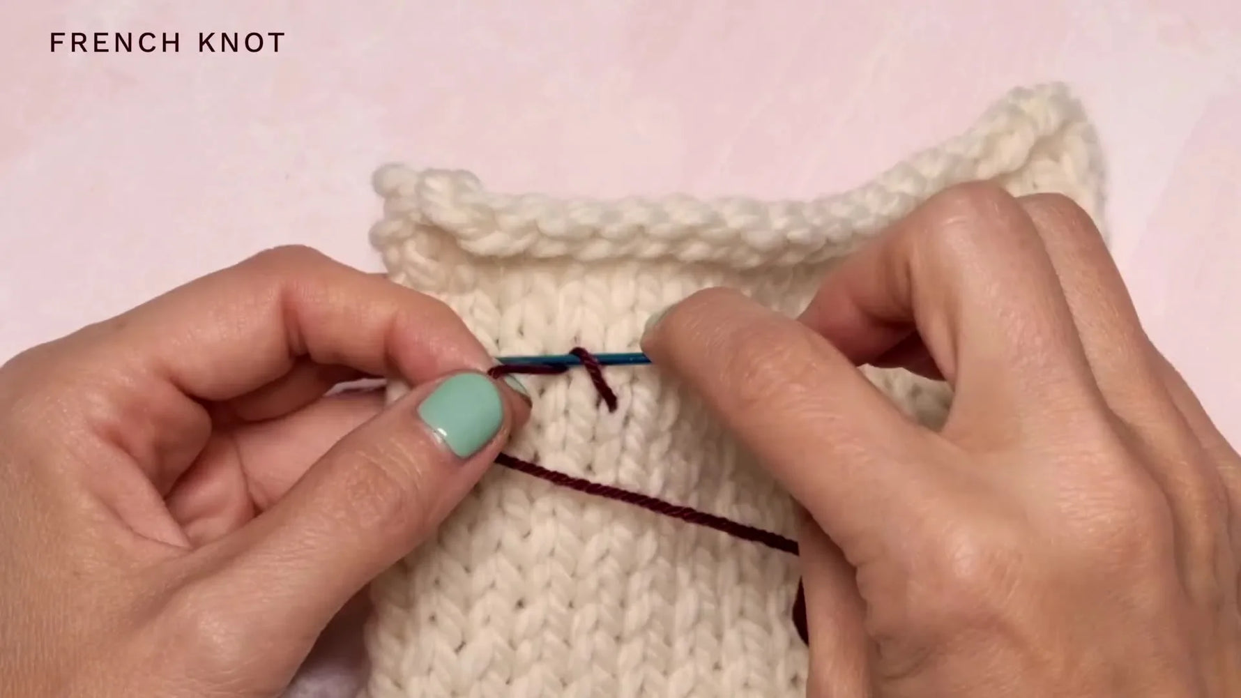Close-up of hands wrapping the dark purple yarn twice around the darning needle, forming the basis of a French knot.