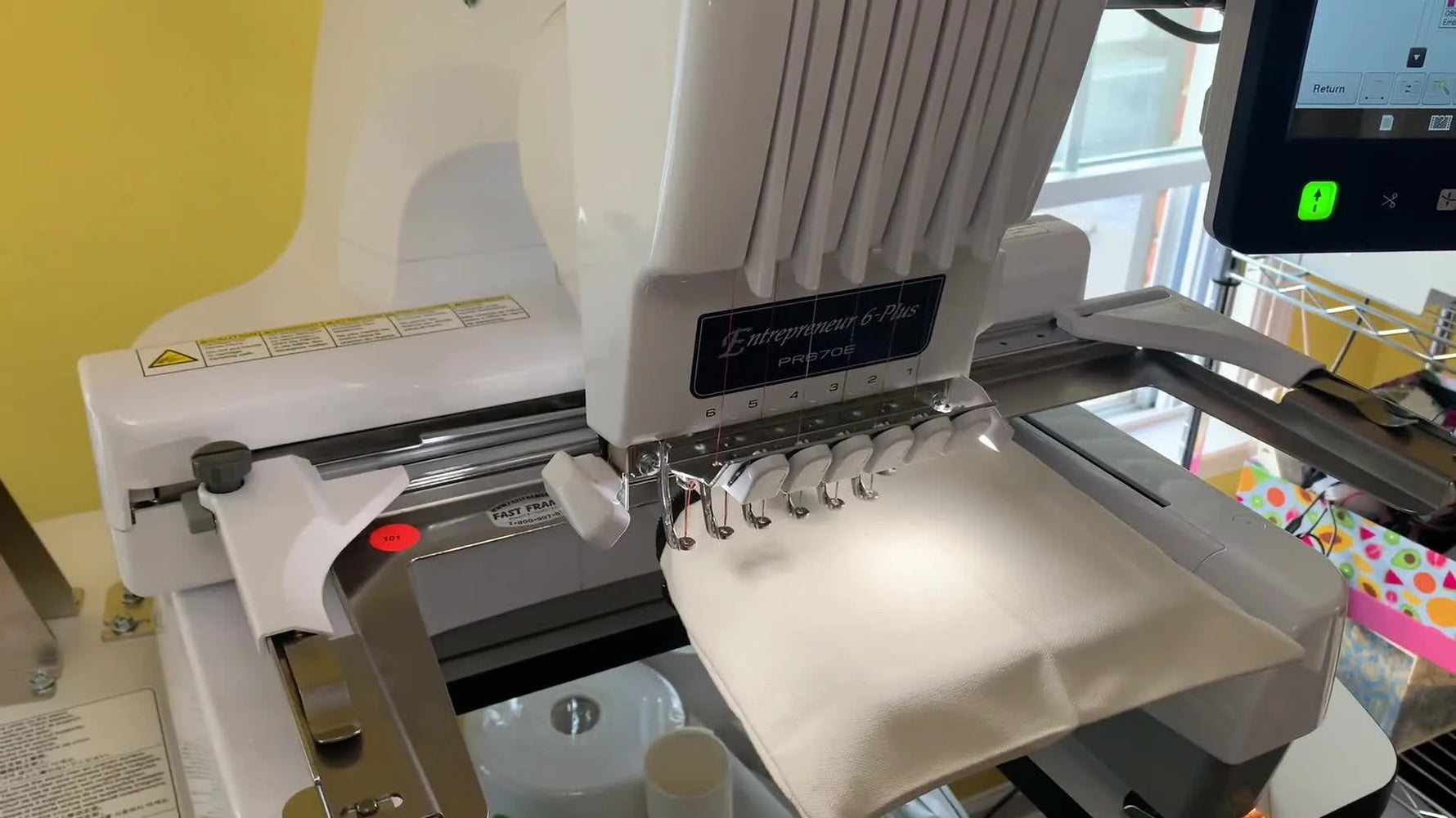 A close-up of a multi-needle embroidery machine stitching a teal letter onto a canvas makeup bag.