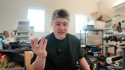 A shop owner holds up three fingers in front of commercial embroidery machines while explaining why one machine can’t scale.