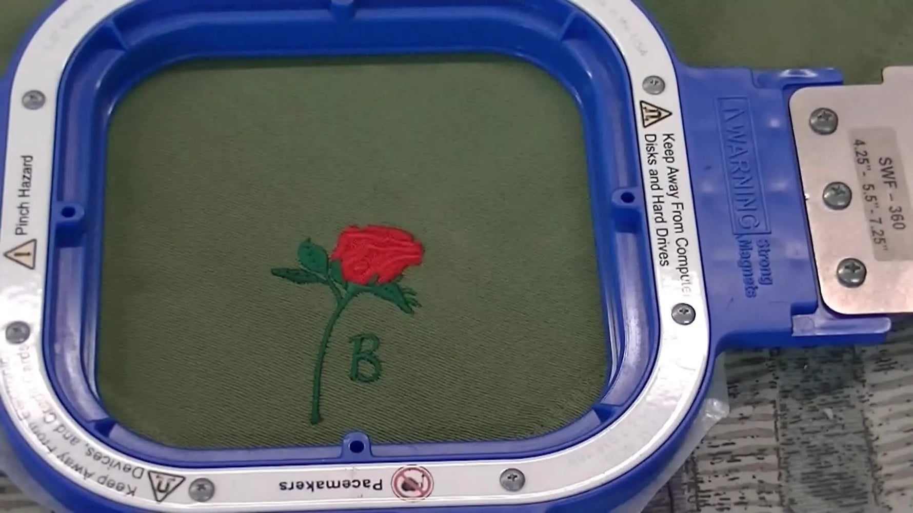 A finished green cotton napkin sits in a blue magnetic hoop, showing a tight rose design with a monogram letter for a clean, production-ready result.