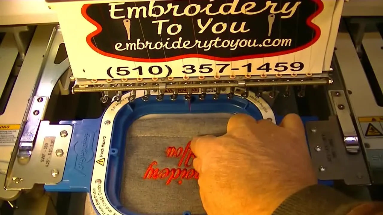 A hand points to the hood hem to show how close the finished embroidery sits to the front edge for a clean, intentional placement.