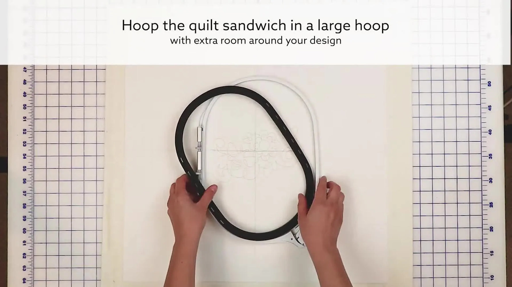 Hands press a large oval hoop over a thick quilt sandwich with a placement template centered for end-to-end quilting alignment.