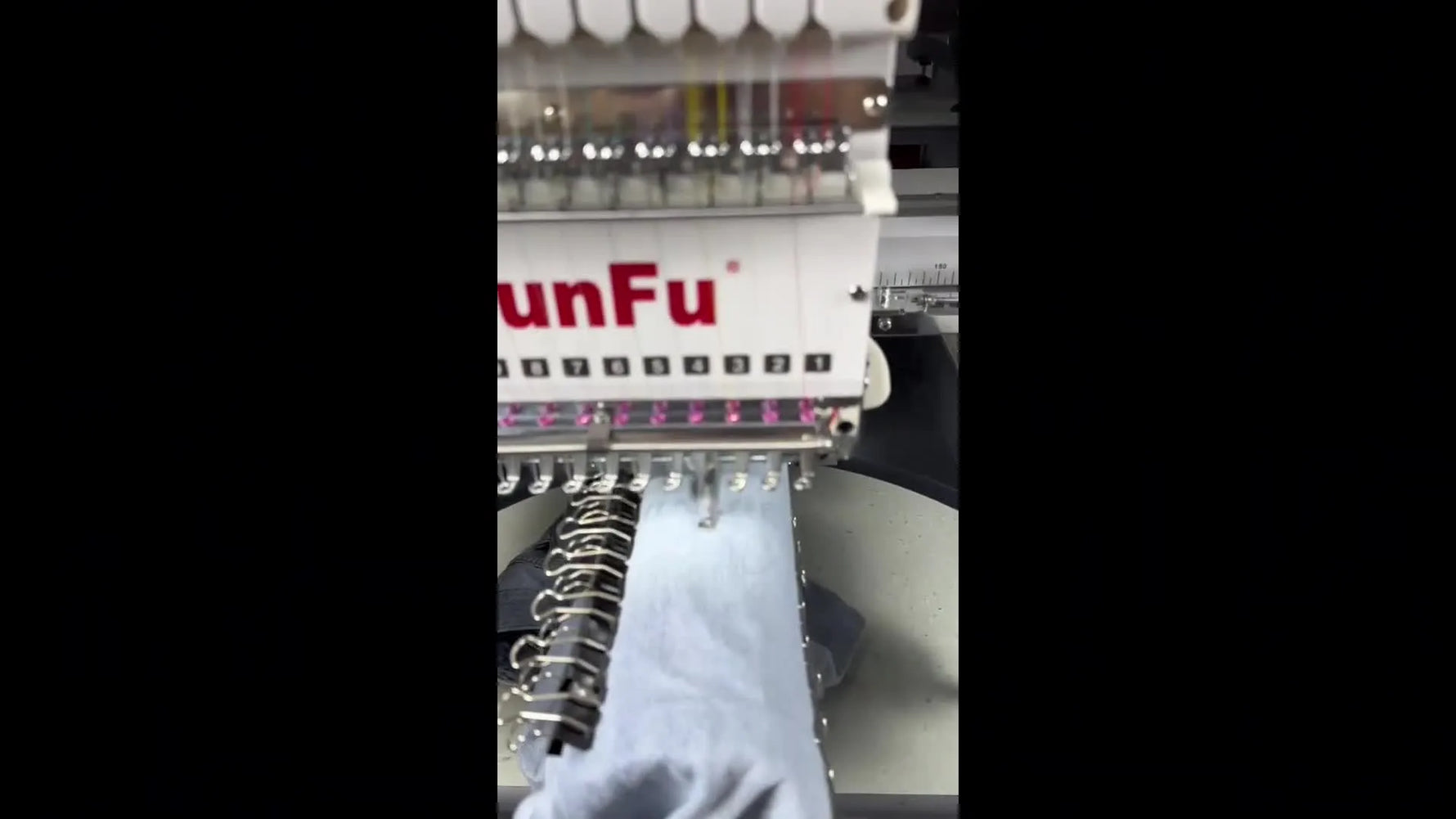 A close-up of the YunFu multi-needle head stitching a purple design on a denim pant leg mounted on the pant device with binder clips visible underneath.
