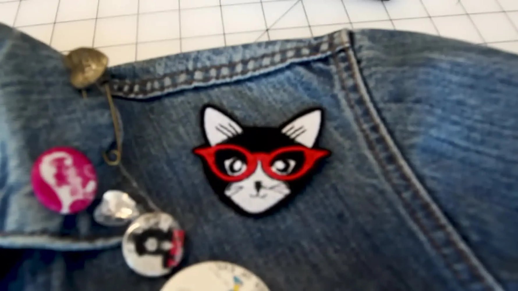 Close-up of a black cat patch with red glasses pinned onto a denim jacket shoulder.