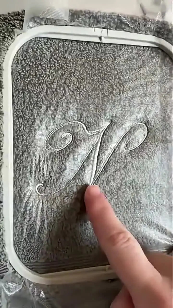 A close-up of a clean, crisp embroidered letter sitting on top of a towel’s pile with the topper still visible.