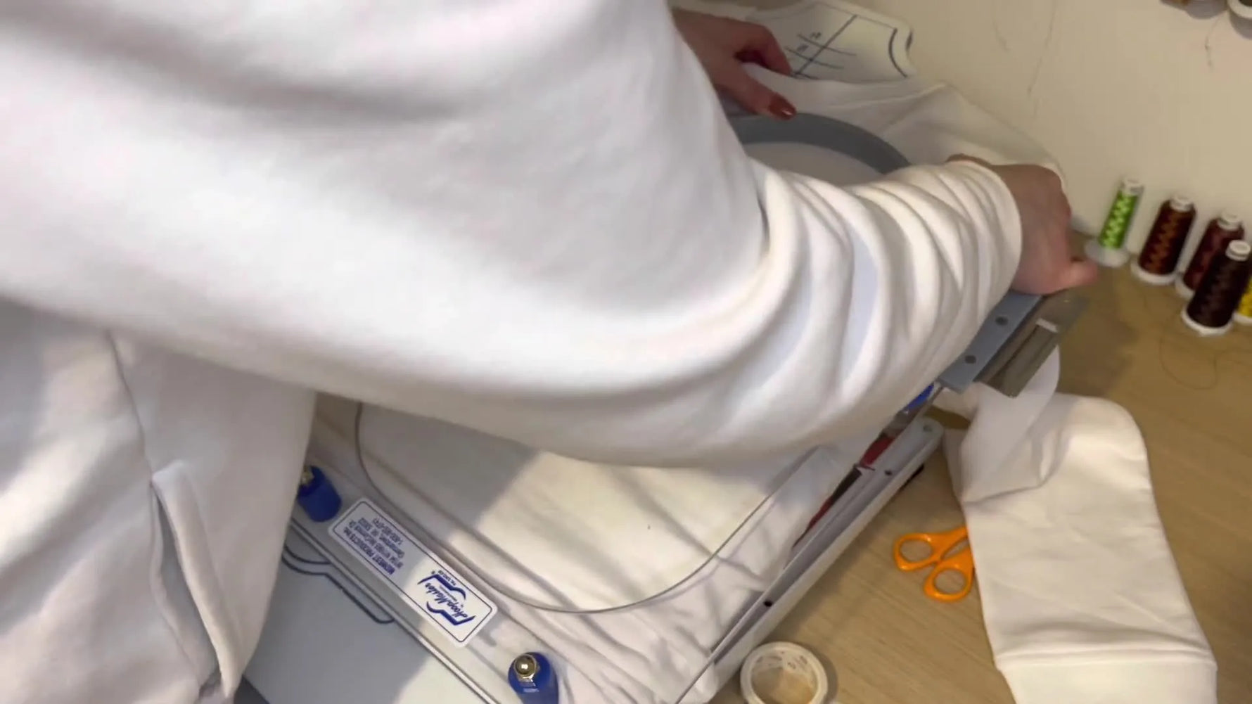Hands press the top hoop down on a Hoop Master fixture to lock a sweater in perfect left-chest position.