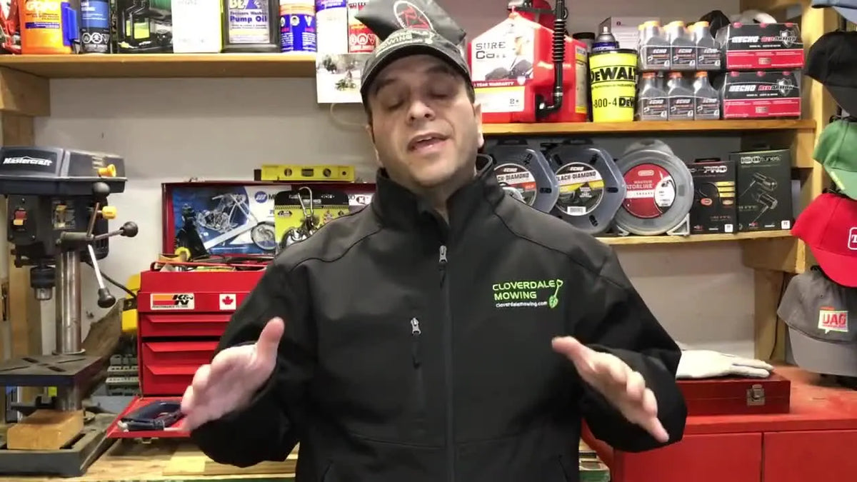 Man in a black jacket with a green lawn care logo, standing in a workshop.