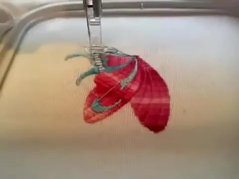A close-up mid-stitch view of the butterfly embroidery showing teal detailing layered over the pink wing fill inside the hoop.