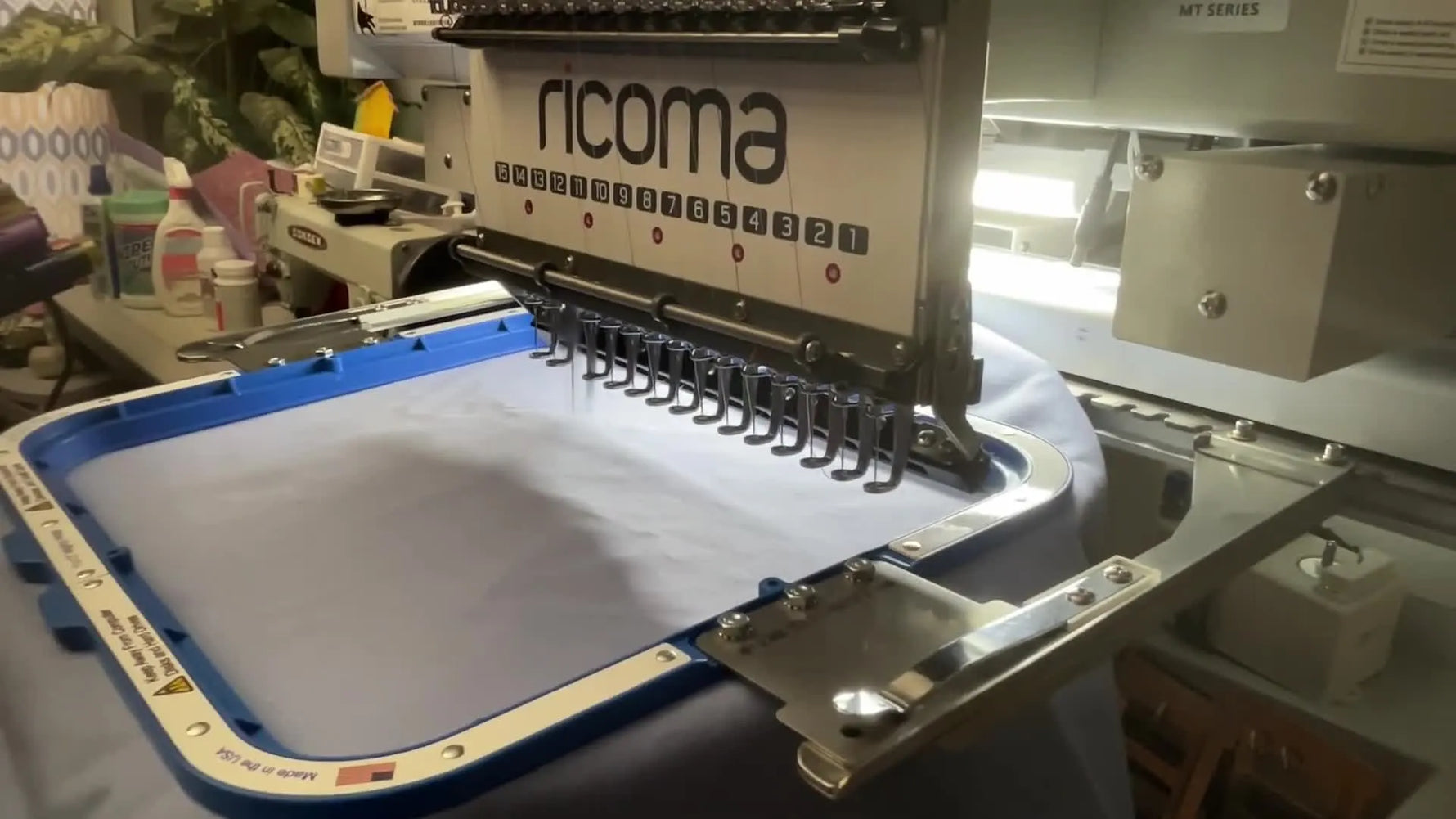 Close-up action shot of the Ricoma MT-1501 stitching the grey outline of the cross on the blue banner fabric inside a magnetic hoop.