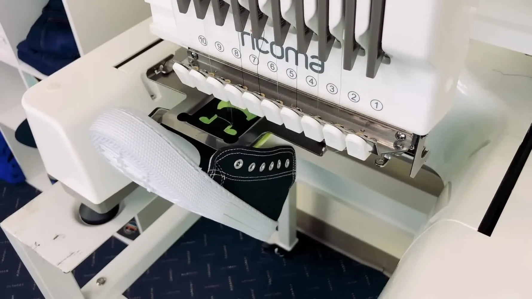 A close-up of the needle stitching a green monster design onto a black canvas shoe tongue held securely in a clamp.