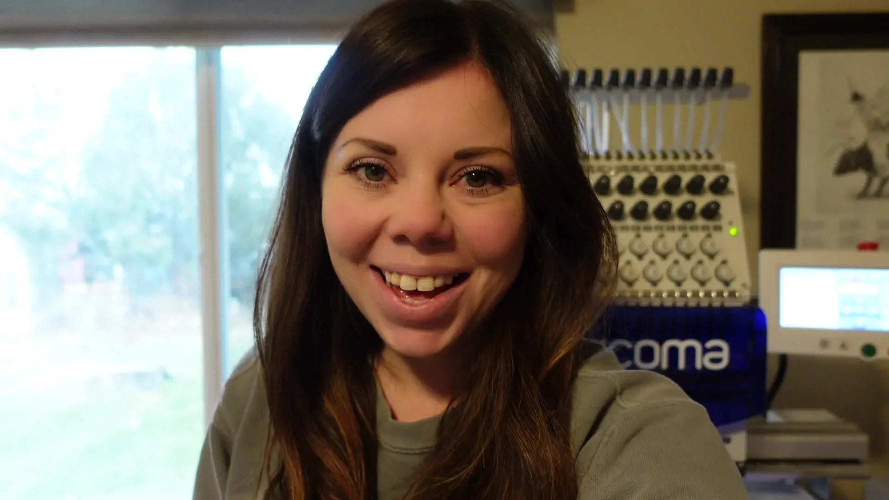 Woman smiling at camera with a Ricoma embroidery machine visible in the background, signaling the start of her VLOGMAS Day 2.