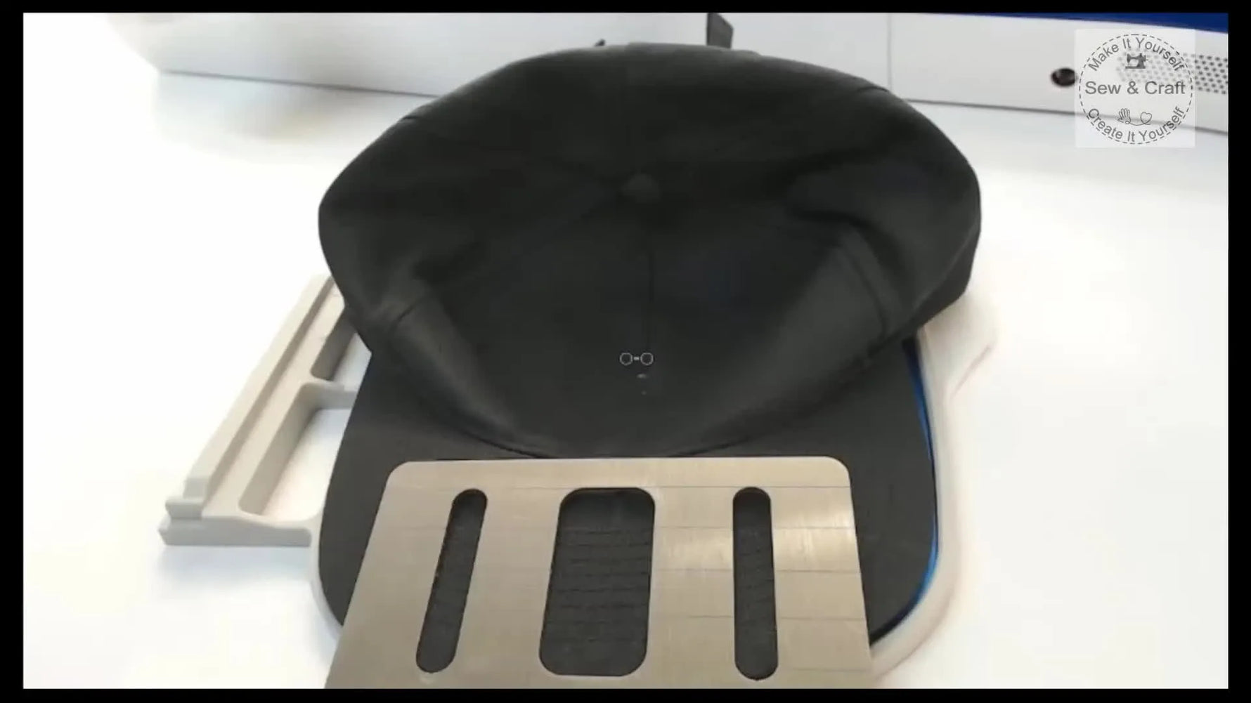 A black baseball cap is mounted flat and secure on an aluminium cap frame insert, ready to stitch on a single-needle embroidery machine.