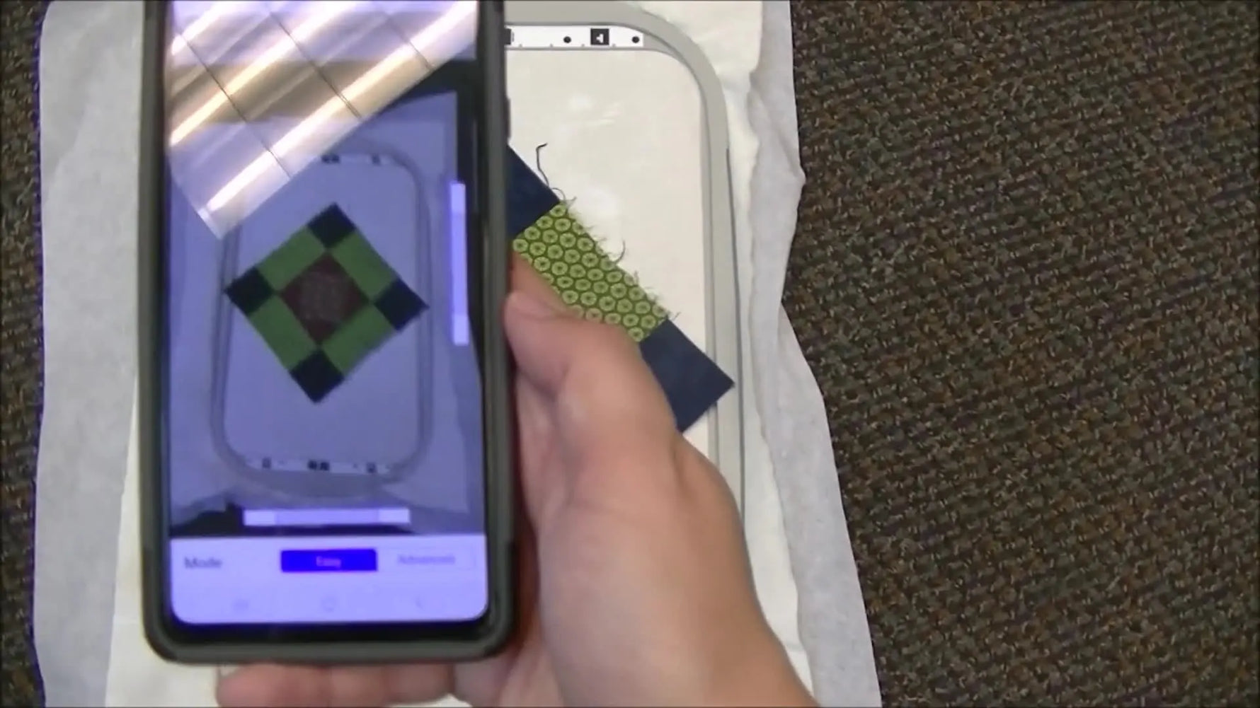 A phone held parallel over a hooped quilt block shows the IQ Intuition AR grid locking onto the hoop’s registration marks for accurate placement.