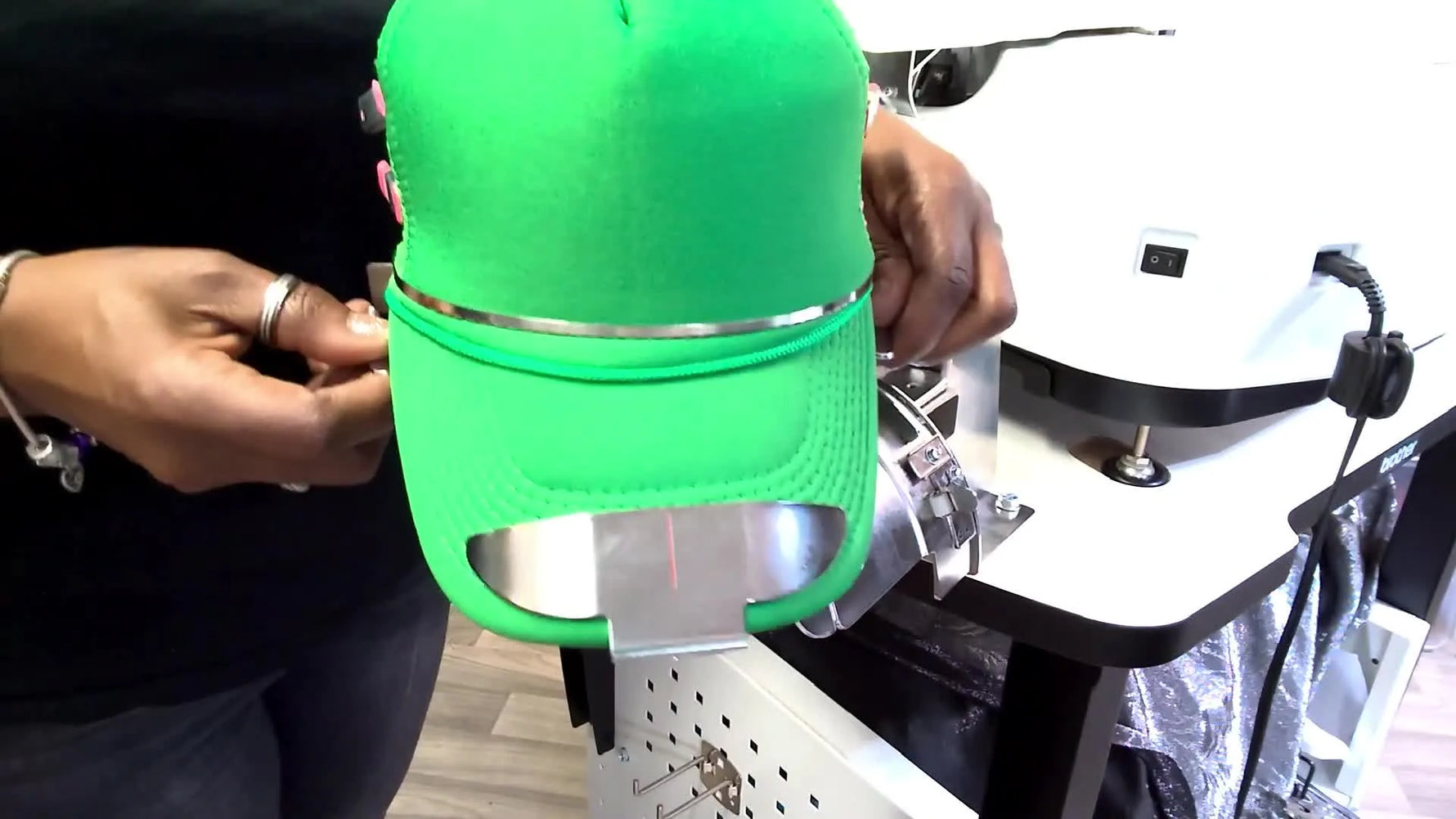 A fully hooped foam trucker hat is held up off the station, showing a flat, tight front panel with stabilizer positioned behind the embroidery area.