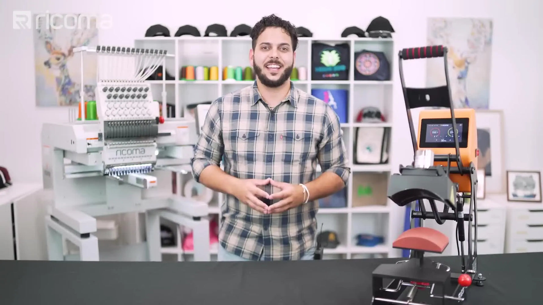 Host standing in front of Ricoma embroidery machine and cap heat press, gesturing.