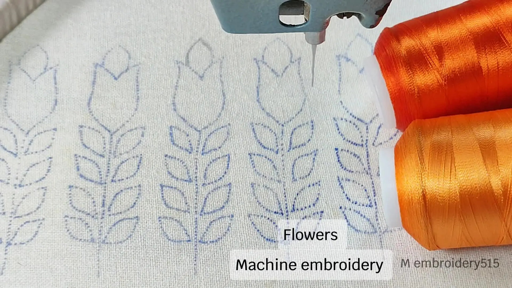 An overhead shot showing embroidery machine needle hovering over a fabric with drawn floral patterns, with orange thread spools nearby.