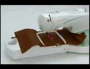 A Singer Futura stitches a pink flower inside a hoop on brown fabric, showing the machine running in embroidery mode.