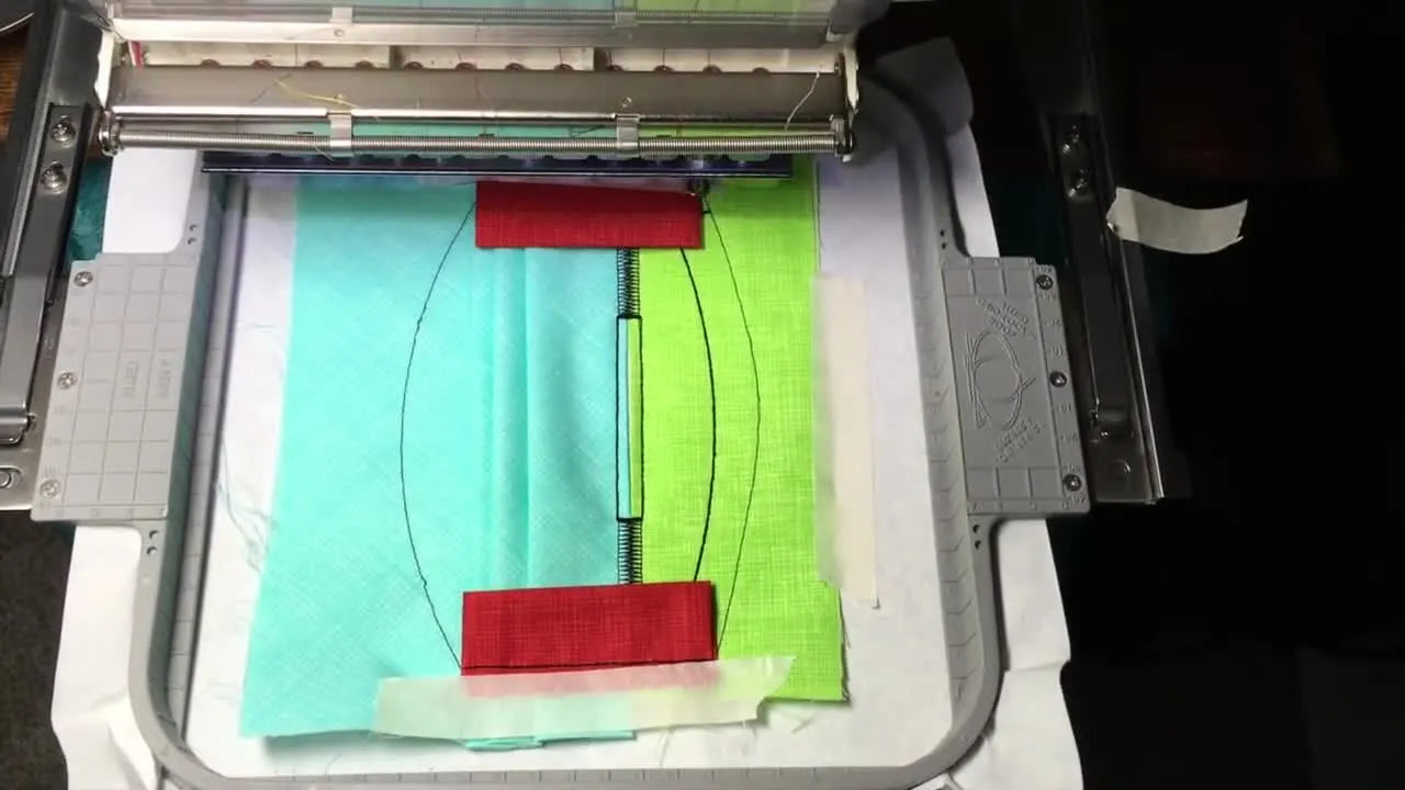 A close-up screen capture of the embroidery machine stitching the side casing pieces while the layered mask fabrics are secured in the hoop.