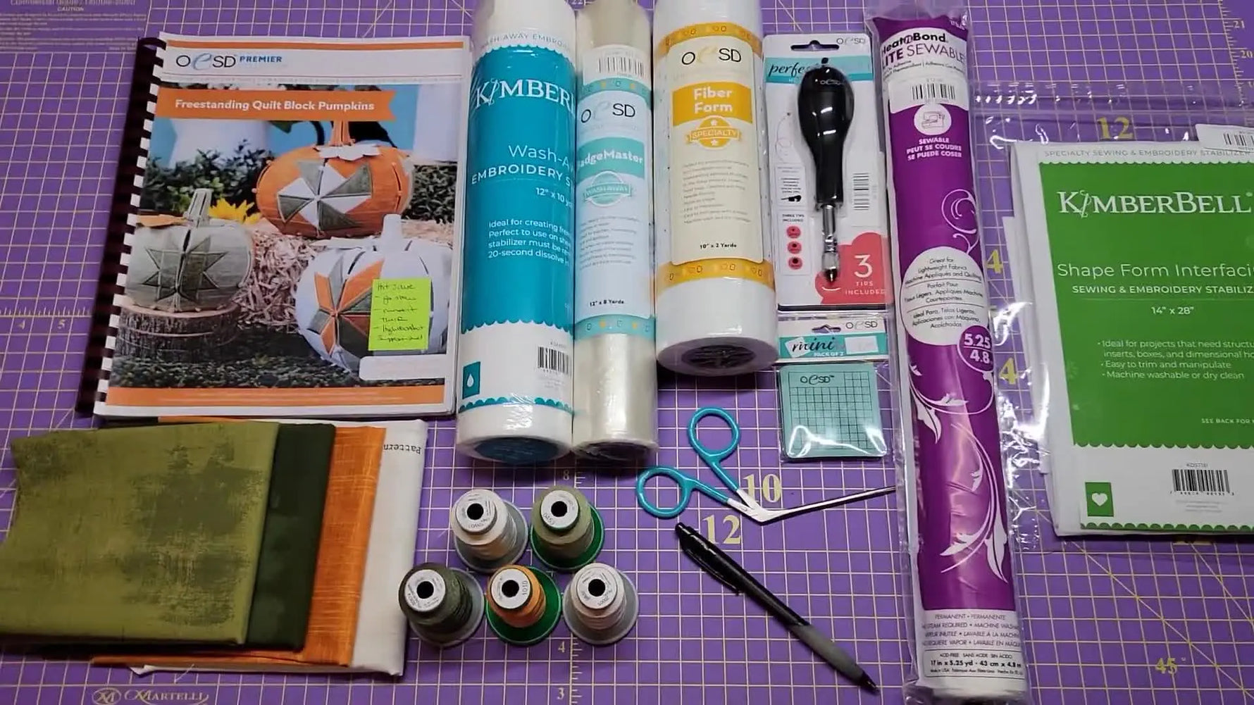 An overhead layout of the OESD pumpkin pattern, stabilizers, fabrics, threads, and specialty tools arranged on a cutting mat before stitching begins.