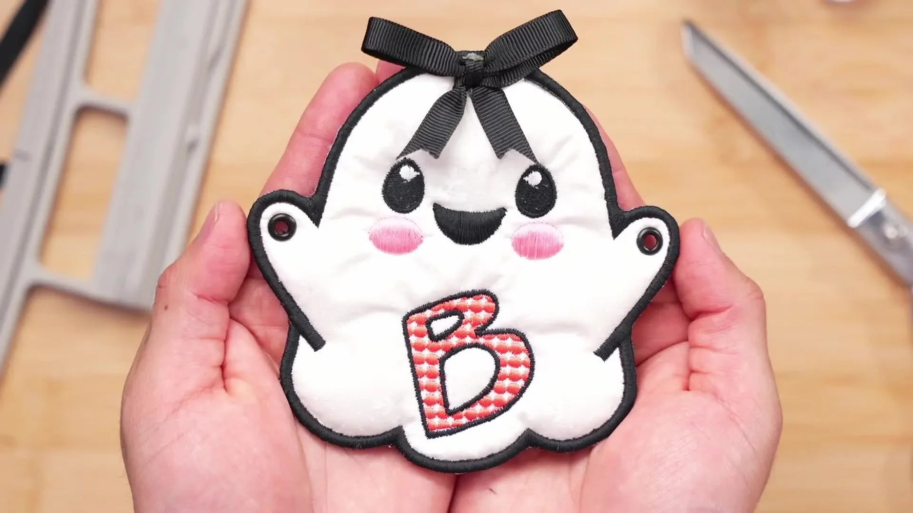 A finished ghost bunting spelling “BOO” is held up on a black ribbon, showing crisp satin borders and clean in-the-hoop construction.
