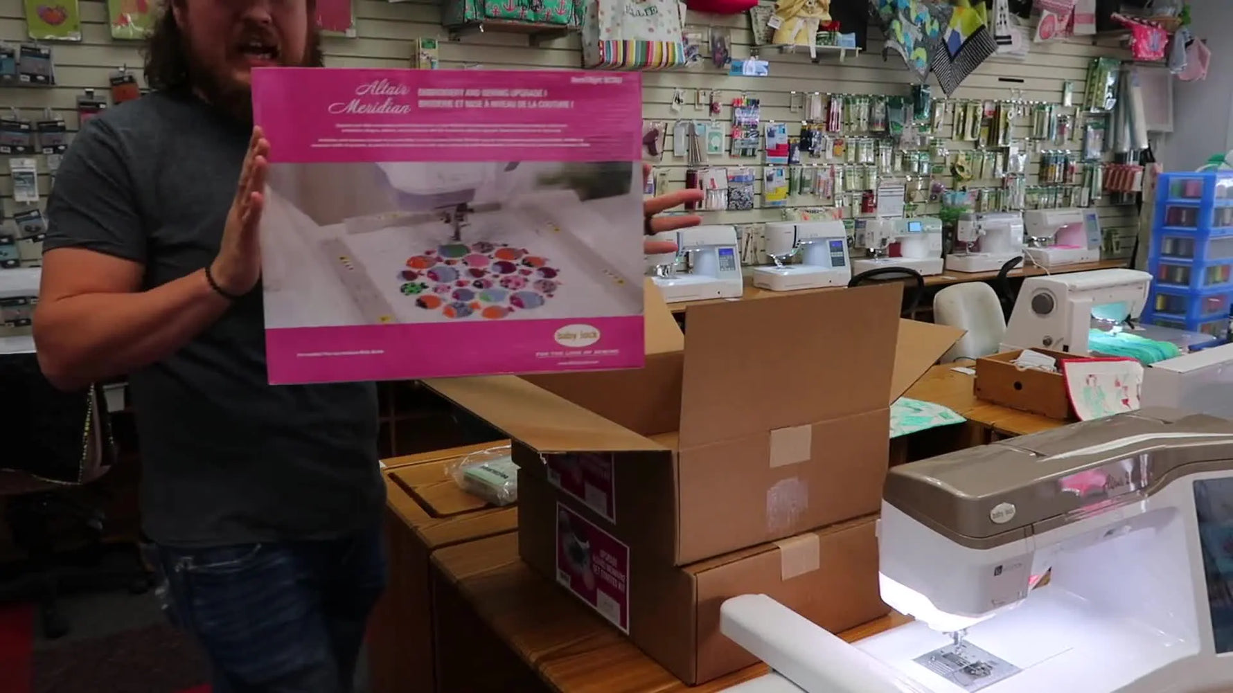 Jordan holds the large pink Baby Lock Altair/Meridian upgrade kit box in the shop, setting up a clear overview of what the upgrade includes.