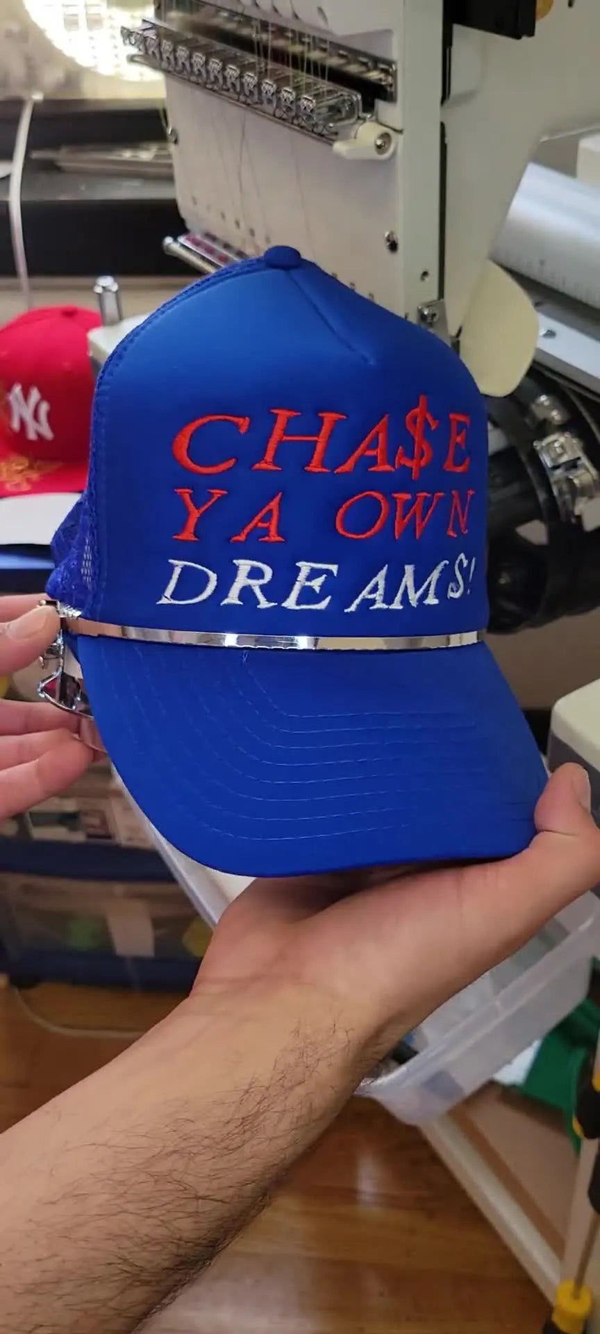 A finished blue foam-front trucker hat is held close to the camera, showing bold stitched lettering and the final texture.