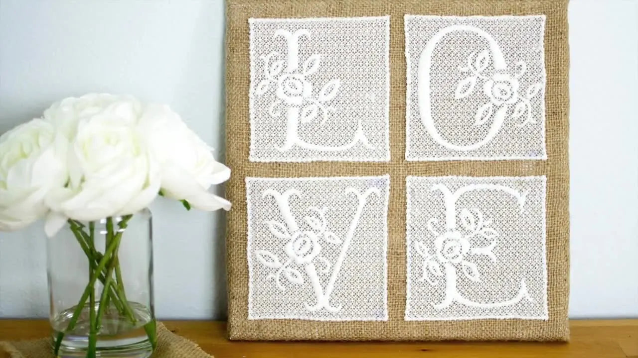 A finished burlap-wrapped canvas displays freestanding lace letters spelling “LOVE,” styled beside white roses.