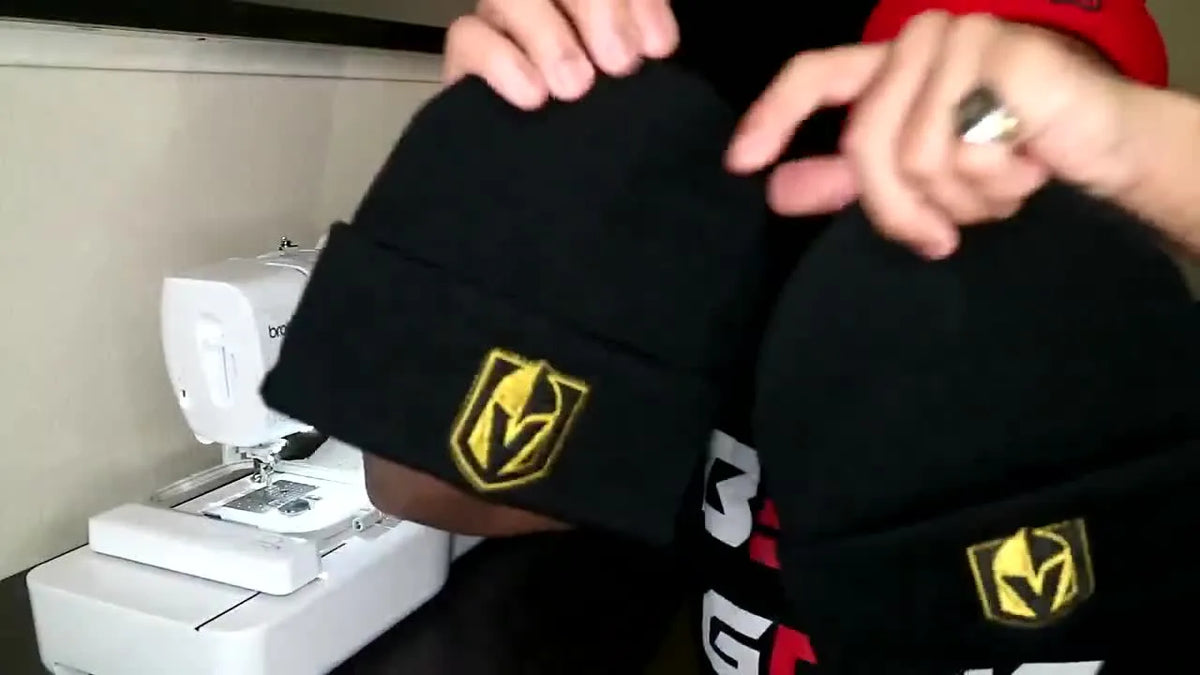 Man proudly holding up two black beanies with identical gold and black shield logos embroidered on them.