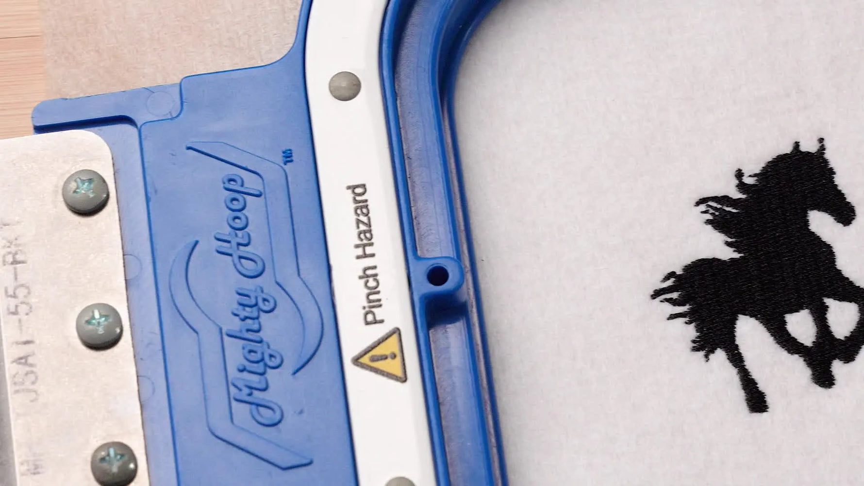 A close-up of a blue magnetic hoop holding white fabric with two stitched horse designs, showing the difference between a flat fill and a textured satin-angle version.