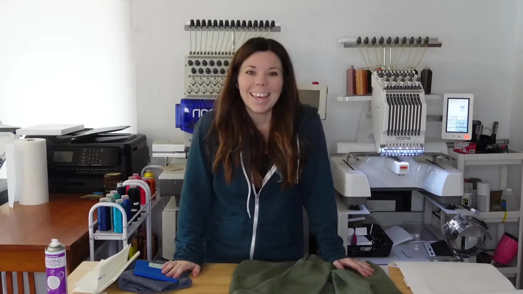Woman in teal hoodie standing in front of two white embroidery machines, showcasing a green sweater.