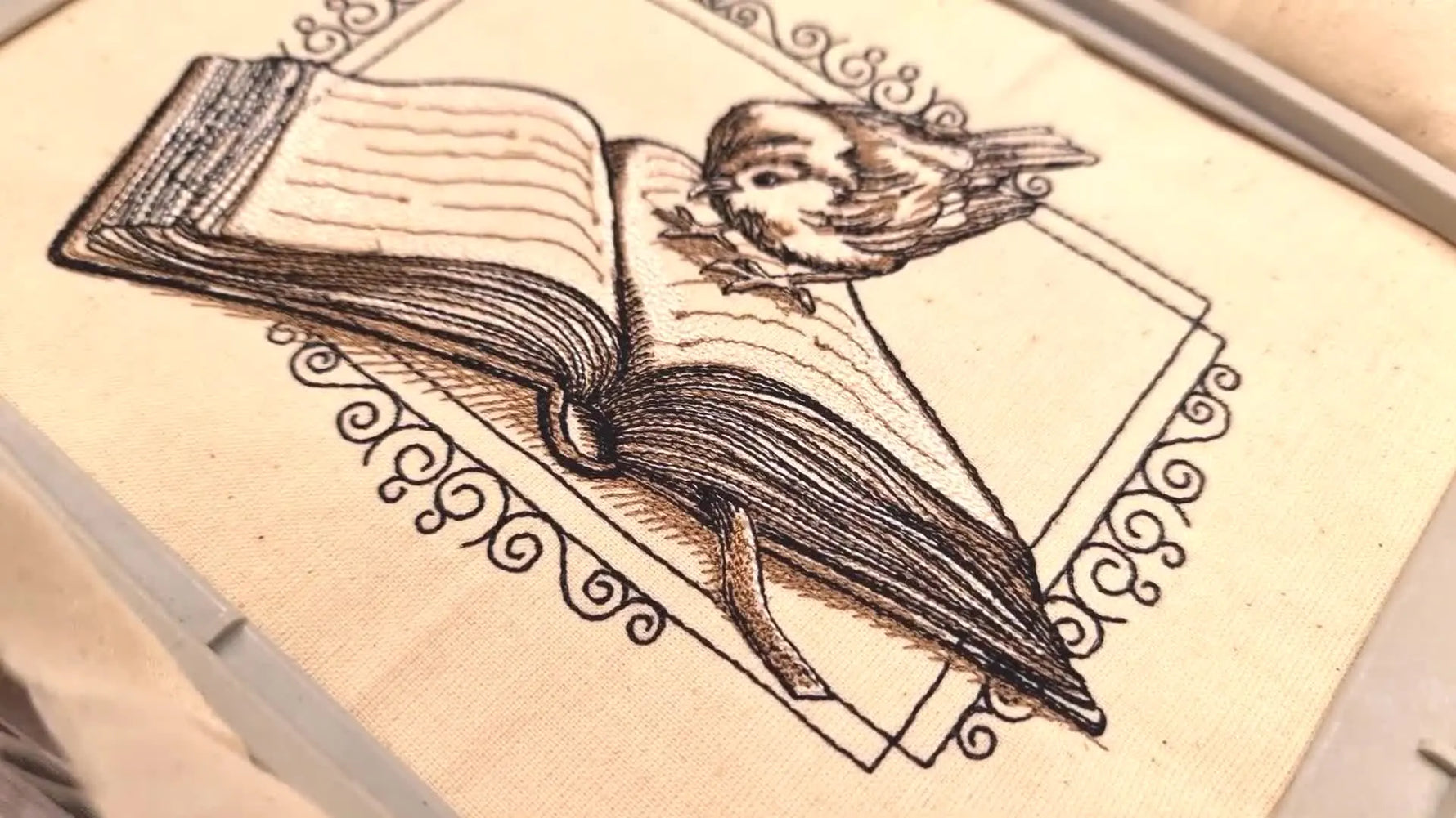 Close-up of the finished embroidered design on fabric, showing a bird on a book within a decorative frame.