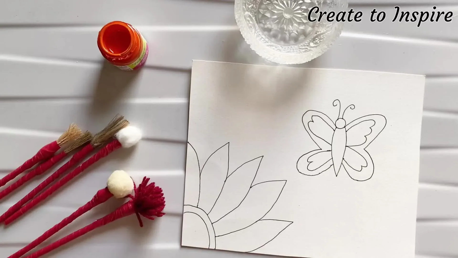 A hand holding a paintbrush, painting a flower, with several DIY brushes and paints laid out.