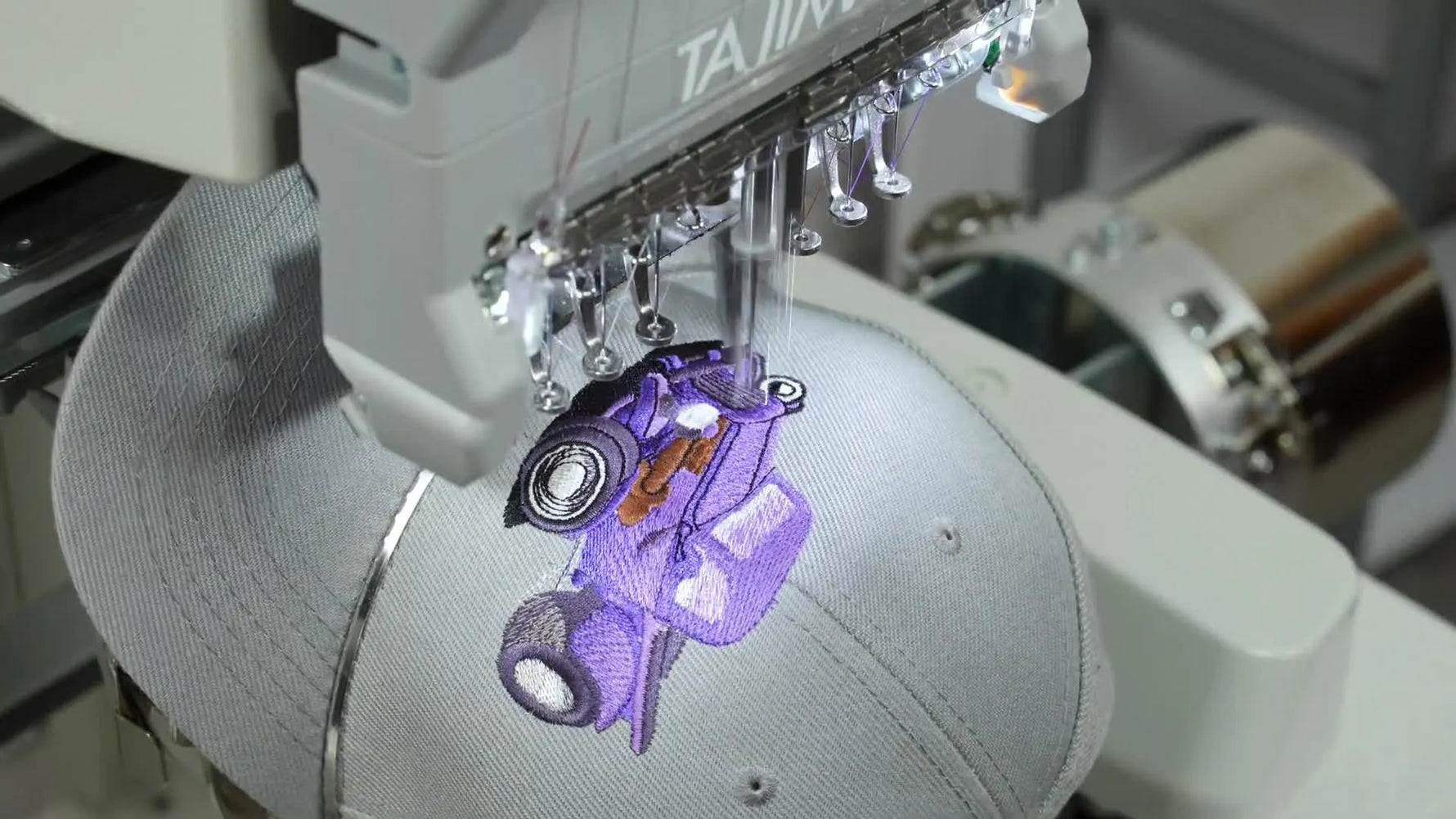 Close-up of Tajima Sai embroidering a purple truck design on a grey hat.
