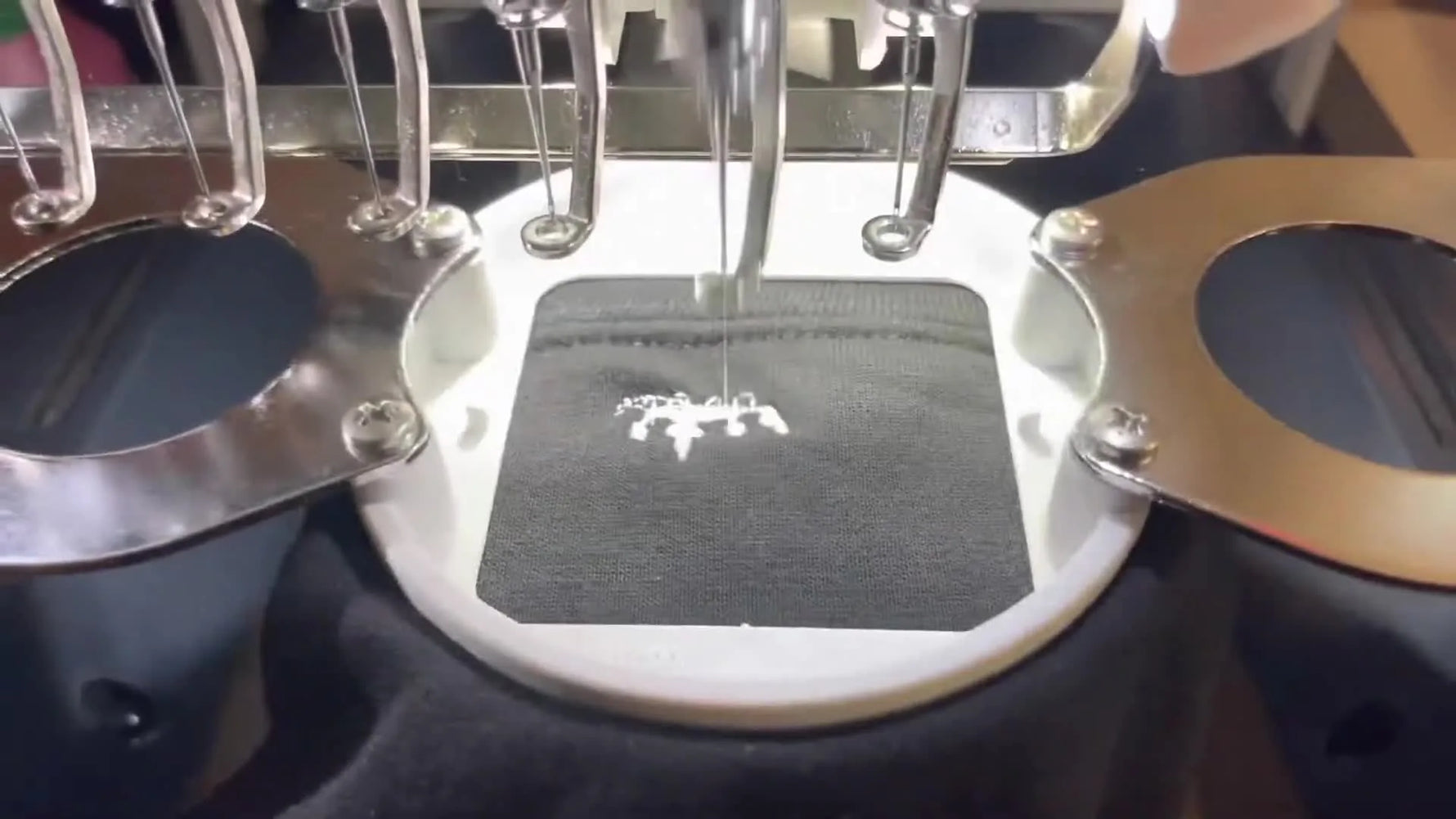 A close-up action shot of a Janome multi-needle machine stitching a small white sleeve logo on black hoodie fabric in a 2x2 hoop.