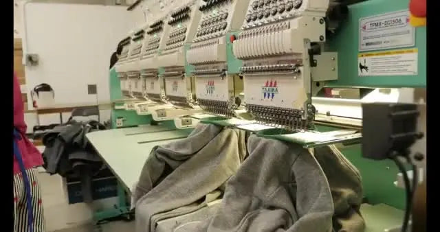 A Tajima multi-head embroidery machine runs a production line on grey garments, illustrating why DST became the practical standard for commercial embroidery.