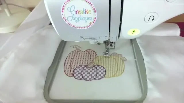An overhead view of the finished metallic pumpkin embroidery stitched on a hooped white tea towel, showing the shimmer and clean placement above the ribbon border.