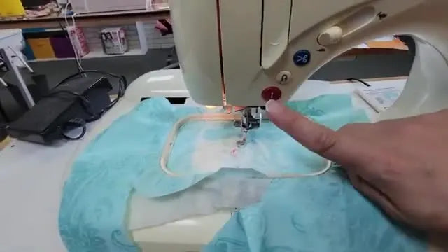 A close-up of a hand pressing the red Start/Stop button as the Singer Quantum XL-100 begins an embroidery stitch-out on hooped fabric.